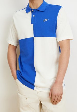 Kortærmet polo med blå og hvide farveblokke, Nike-logo på brystet, båret af en person med den ene hånd i lommen.