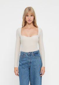 Blonde Frau, die ein beiges Langarm-Oberteil mit quadratischem Ausschnitt und hochgeschnittene blaue Jeans trägt, steht vor einem schlichten weißen Hintergrund.