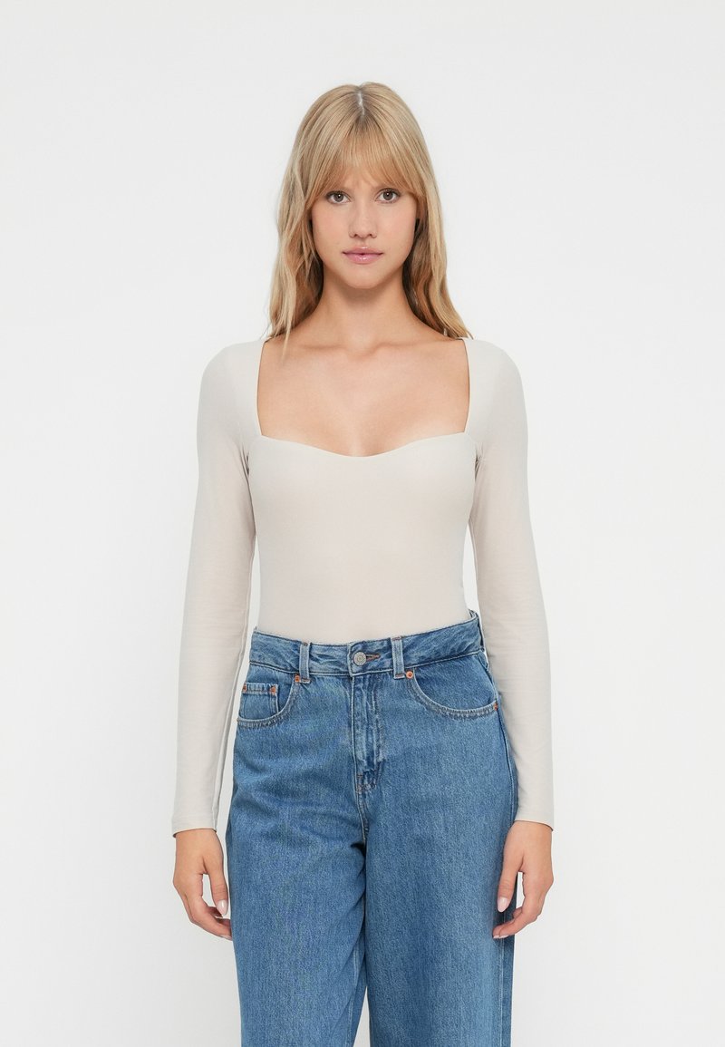 Blonde Frau, die ein beiges Langarm-Oberteil mit quadratischem Ausschnitt und hochgeschnittene blaue Jeans trägt, steht vor einem schlichten weißen Hintergrund.