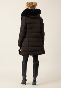 Schwarze Daunenjacke mit einem strapazierfähigen Fellkragen, Taillengürtel und gestepptem Design, getragen über figurschmeichelnden schwarzen Hosen und eleganten kniehohen Stiefeln.
