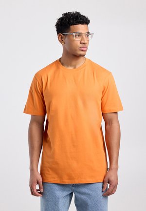 Junger Mann mit kurzen lockigen Haaren, klaren Brillen, orangefarbenem T-Shirt und hellblauen Jeans, schaut nach rechts vor einem einfarbigen Hintergrund.