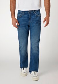 Mėlynos džinsinės kelnės su tiesiu kirpimu, išblukusia tekstūra, priekinėmis kišenėmis ir mygtuko uždarymu, derinamos su baltais sportiniais bateliais.