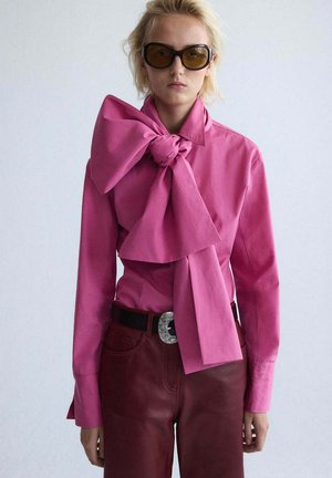 Personne portant de grandes lunettes de soleil foncées, un chemisier rose vif avec un nœud surdimensionné au col, et un pantalon taille haute rouge foncé avec ceinture.