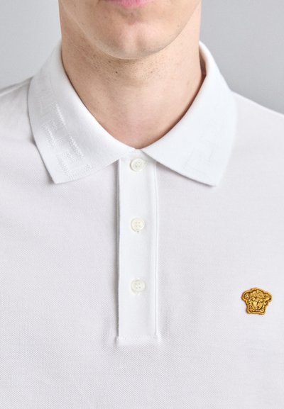 Polo shirt blanche avec un col texturé, trois boutons et un petit logo doré brodé sur la poitrine.