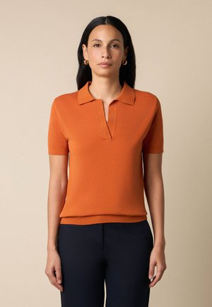 Kobieta o długich ciemnych włosach, nosząca złote kolczyki koła, pomarańczową koszulkę polo z krótkim rękawem i ciemne spodnie na beżowym tle.