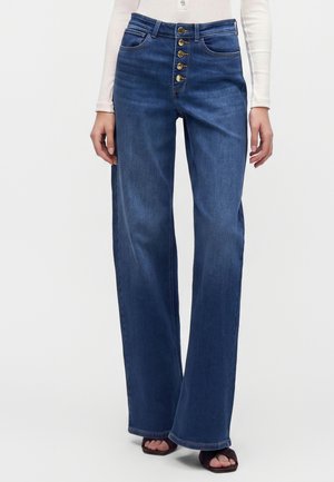 Donna che indossa jeans blu a vita alta a gamba larga con patta con bottoni a vista e una maglia bianca a maniche lunghe, in piedi davanti a uno sfondo neutro.