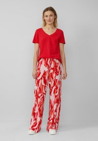 Czerwony t-shirt z dekoltem w serek w połączeniu z luźnymi, plisowanymi spodniami w czerwono-białym wzorze. Model nosi białe trampki, prezentując swobodny wygląd.