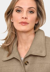 Beige gewatteerde jas met een puntige kraag, voorzien van een grote knoop. Zachte textuur met zichtbare stiksels.