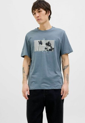 T-shirt en coton bleu clair avec une impression graphique rectangulaire représentant des palmiers et le texte "WORLD ORIGINALS." Manches courtes, col rond.