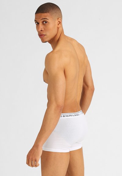 Witte katoenen boxershorts met een zwart logo op de tailleband. Het model staat met een blote torso, iets wegdraaiend, en toont een aansluitende snit.