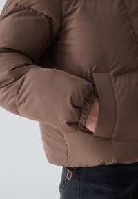 Brązowa kurtka puchowa o gładkim, matowym wykończeniu. Posiada elastyczne mankiety, boczną kieszeń oraz sznurek regulujący na dole. Ciepła, ocieplana konstrukcja.