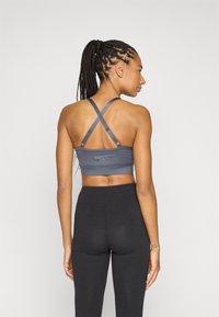 Šedá sportovní podprsenka s překříženými ramínky, s přiléhavým střihem a hladkou texturou. Outfit doplňují černé legíny s vysokým pasem.