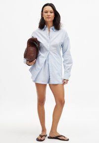 Light blue checked shirt with long sleeves, paired with matching shorts. Holding a woven brown bag. Model wears black flip-flops.