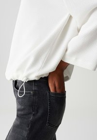 Persoon in een textuurwitte jas met een trekkoord aan de onderkant en zwarte jeans, hand rustend in de achterzak tegen een effen achtergrond.