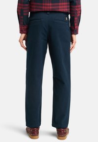Navy blue cotton pants with a straight-leg cut, featuring two back pockets and a small logo tag at the waistband. Worn with brown footwear.