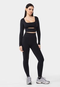 Crna dugih rukava cropped majica s izrezima, u kombinaciji s crnim legging hlačama visokog struka. Model nosi svijetlosive sportske tenisice s tamnim akcentima.