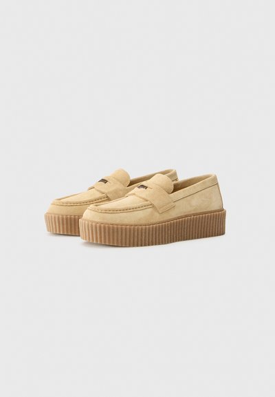 Mocassins en suede beige avec une semelle en caoutchouc épaisse et rainurée, présentant un design cousu et une bande décorative à l'avant. Style classique à enfiler.