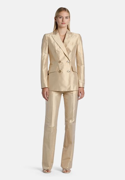 Blonde woman wearing a beige double-breasted blazer and matching straight-leg pants, standing with hands at sides on white background.