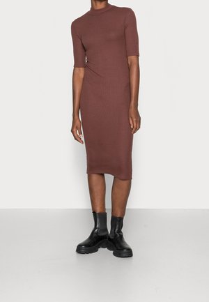 Femme portant une robe midi marron cintrée avec des manches au coude et des bottines noires épaisses, debout devant un fond clair uni.