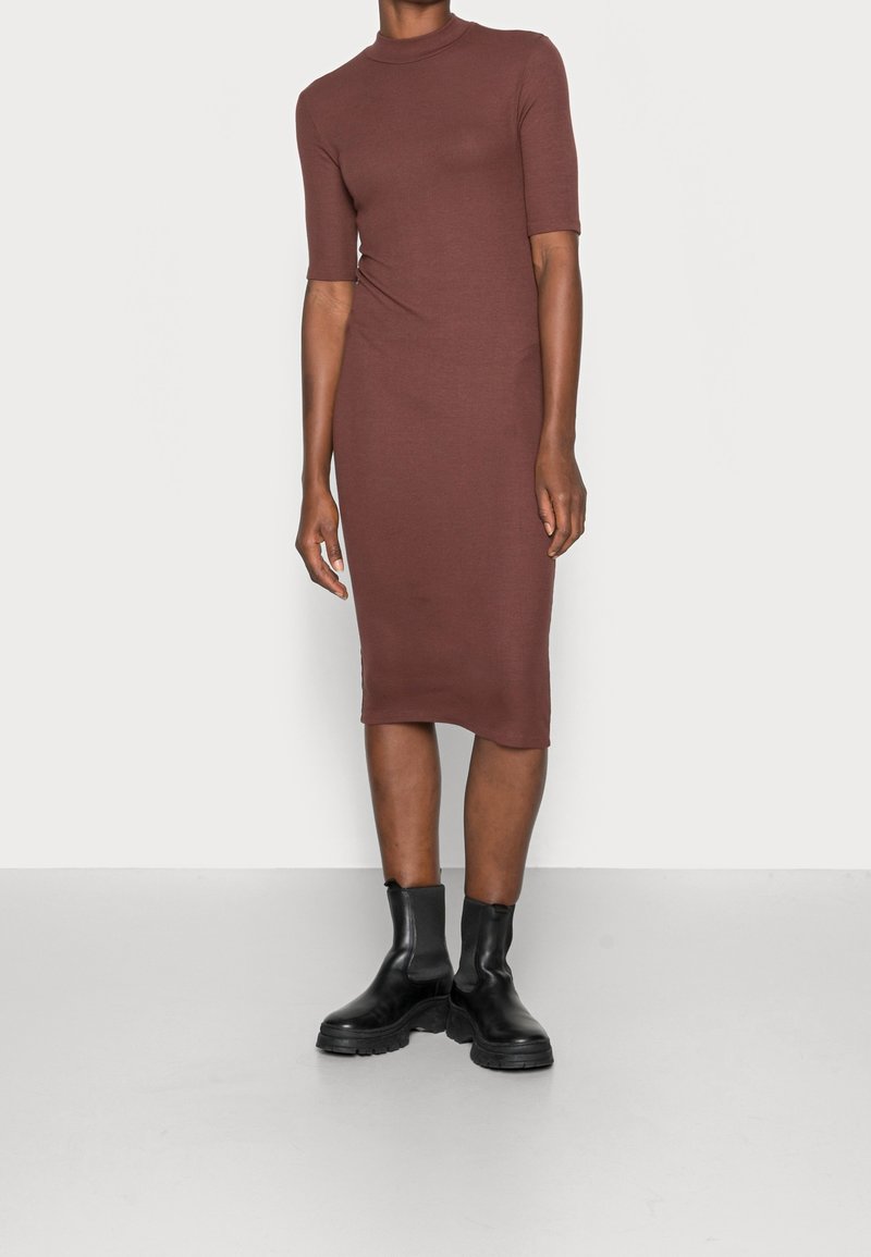 Femme portant une robe midi marron cintrée avec des manches au coude et des bottines noires épaisses, debout devant un fond clair uni.