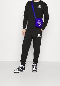 Svart sweatshirt och joggers med vita logotyper, tillsammans med svart och vita sneakers. En liten lila crossbody-väska bärs över kroppen.