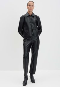 Vrouw in een aansluitende zwarte leren jas, bijpassende leren broek en zwarte hoge enkelboots, staand tegen een lichte achtergrond.