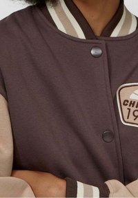 Osoba v hnědo-bežovém varsity bundě s knoflíky a nášivkou s nápisem "CH 19." Ruce jsou založené v pase.
