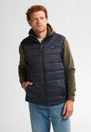 Vest negru cu umplutură, cu închidere cu fermoar, model de cusături verticale și buzunare. Puratat peste un pulover verde cu glugă.