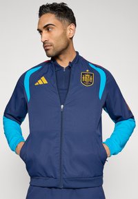 Chaqueta azul marino con cremallera y detalles en turquesa, que presenta un escudo español, logo amarillo de Adidas y puños acanalados. Fabricada con material ligero.