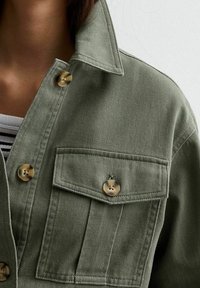 Groene denim jacket met een spitse kraag, twee borstzakken en ronde metalen knopen. De stof heeft een gestructureerde afwerking.