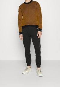 Brązowy sweter z dzianiny z czarnym wykończeniem, w połączeniu z czarnymi spodniami dresowymi z bocznymi paskami. Całość dopełniają beżowe trampki.