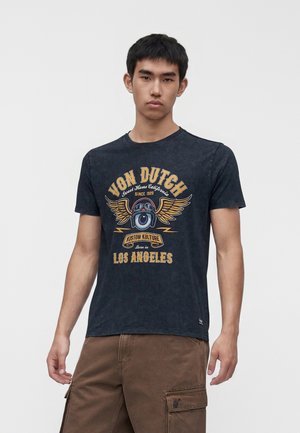 Junger Mann trägt ein schwarzes Von Dutch T-Shirt mit Flügeln, Schutzbrille und dem Schriftzug „Los Angeles“ sowie braune Cargohosen.