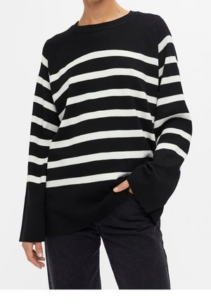 Personne portant un pull à rayures horizontales noires et blanches à manches longues amples et un pantalon noir, main dans la poche.