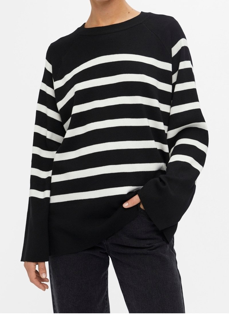 Personne portant un pull à rayures horizontales noires et blanches à manches longues amples et un pantalon noir, main dans la poche.