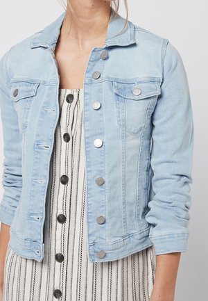 Femme portant une veste en denim bleu clair avec des boutons en métal sur une robe blanche à rayures noires verticales et grands boutons noirs.