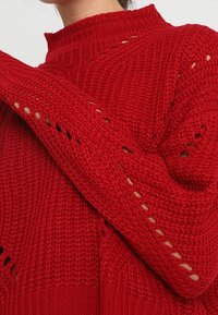 Pull en tricot rouge avec un col montant, un tissu texturé et des motifs ajourés le long des manches pour un détail supplémentaire.