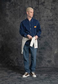 Camicia di jeans blu con tasca a toppa e etichetta arancione, abbinata a jeans blu e sneakers marroni. Un felpa bianca è legata in vita.