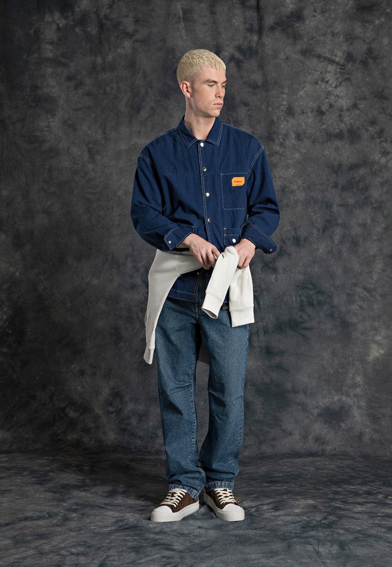 Camisa de ganga azul com um bolso de chapa e etiqueta laranja, combinada com jeans azuis e ténis castanhos. Um sweatshirt branco está atado à cintura.