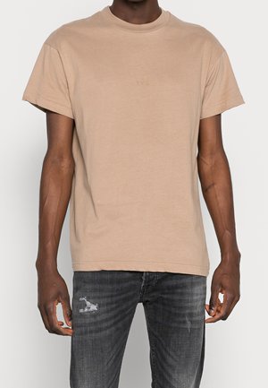 Beige bomulds t-shirt med korte ærmer, rund hals, prydet med "YPS" trykt i en diskret skrifttype. Båret med slidte mørke jeans.