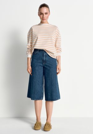 Frau steht und trägt ein langärmeliges Oberteil mit Pfirsich-Streifen, knieumspielende, weite Jeansshorts und hellbraune Mokassins vor einem einfarbigen Hintergrund.