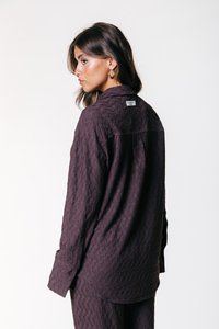 Camicetta viola scuro, testurizzata, con una vestibilità ampia, polsini con bottoni e un'etichetta sul retro. Presenta un sottile motivo geometrico.