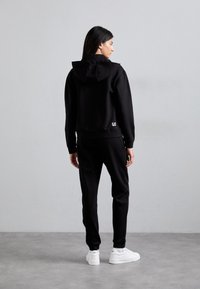 Zwarte hoodie met een capuchon, elastische manchetten en een logo-detail aan de zoom, gecombineerd met bijpassende zwarte broek en witte sneakers.