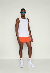 Muž v bielej bezrukávovej košeli, jasne oranžových šortkách, bielych teniskách, bielych ponožkách Nike a reflexných modrých športových slnečných okuliaroch stojí proti jednolitému pozadiu.