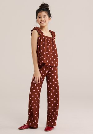 Niña sonriente con moño, vestida con top marrón sin mangas y pantalones con lunares rosa y bailarinas rojas, de pie sobre un fondo beige.