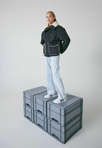 Schwarze gepolsterte Jacke mit einem cremefarbenen Kunstfellkragen, über einem hellblauen Denim-Outfit getragen, steht auf drei grauen Aufbewahrungsboxen.