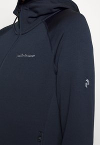 Sweatshirt à capuche de couleur marine en tissu doux, avec une poche zippée et un logo accentué sur la poitrine et la manche. Texture lisse, design ajusté.