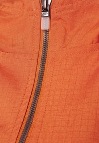 Orange tyg med ett texturerat mönster, med en silvrig metalldragkedja med en logotapp längs mitten.