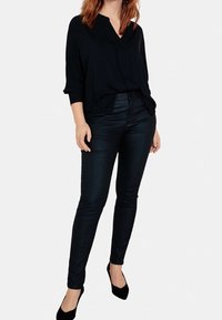 Schwarze, lässig geschnittene Bluse mit V-Ausschnitt und Dreiviertelärmeln; kombiniert mit schwarzen, eng anliegenden Hosen und schwarzen High Heels.