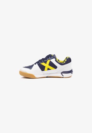 Zapatilla deportiva de corte bajo con malla blanca y azul marino, detalles amarillos, suela de goma y cordones, vista desde el lado exterior sobre fondo blanco.