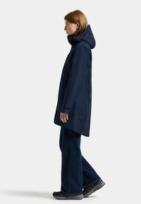 Marinblå vattentät jacka med huva, med rak skärning, sidofickor och justerbara ärmslut. Kombinerad med mörka vida jeans och skor.